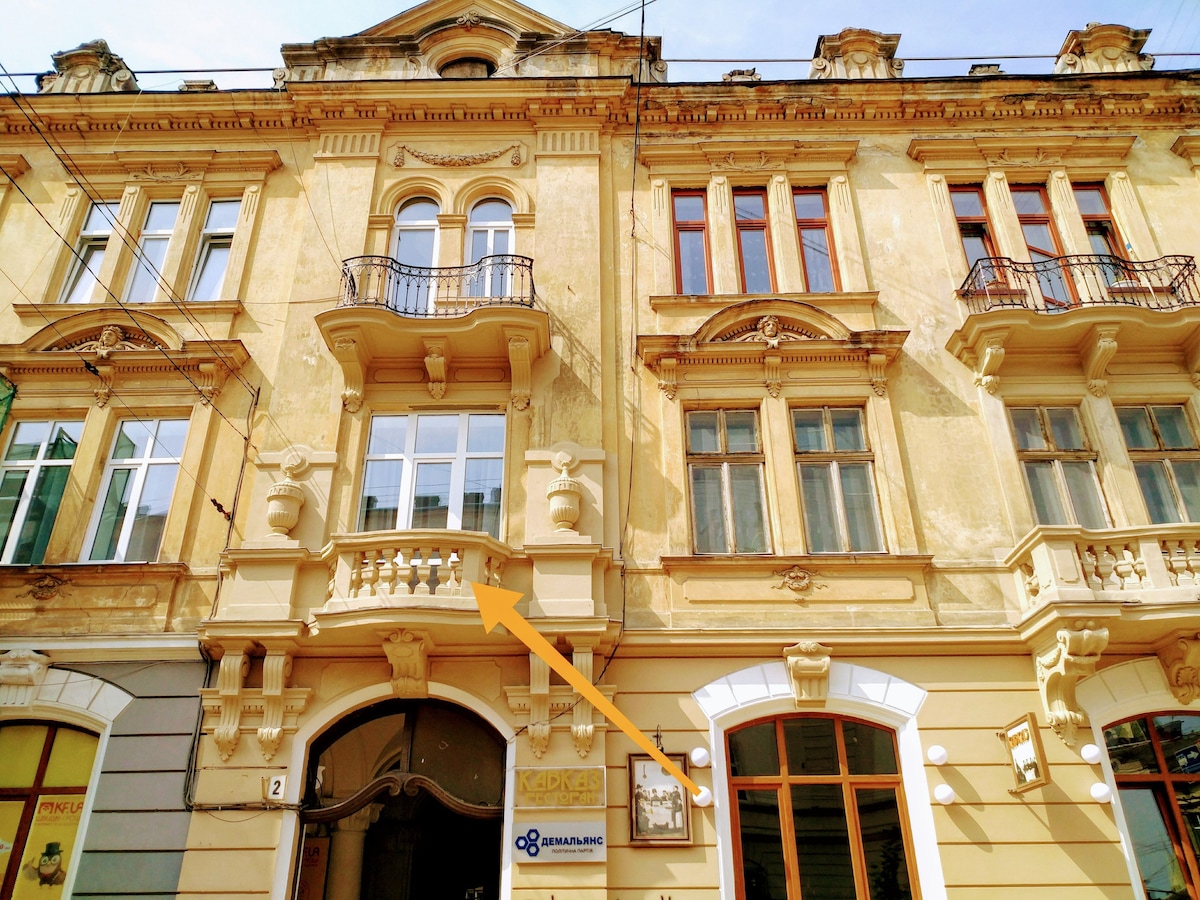 The exterior of a historic building displays ornate architectural details and balcony railings. Large windows reflect the surrounding environment. The façade features warm tones with decorative moldings and identifies the entrance below, ensuring easy access for guests.