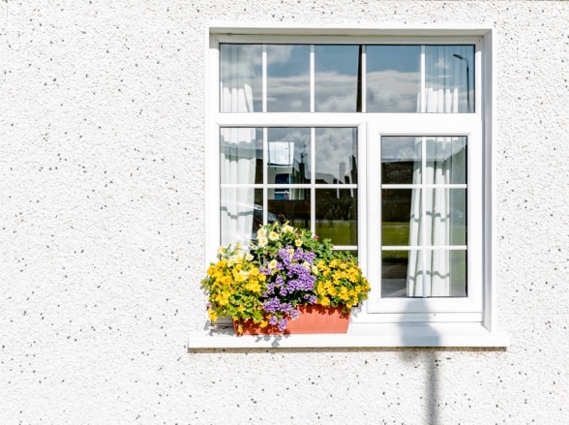 A bright window features a white frame divided into six panes, adorned with sheer white curtains. Below, a planter holds vibrant flowers in shades of yellow and purple, creating a welcoming exterior that enhances the home’s charm.