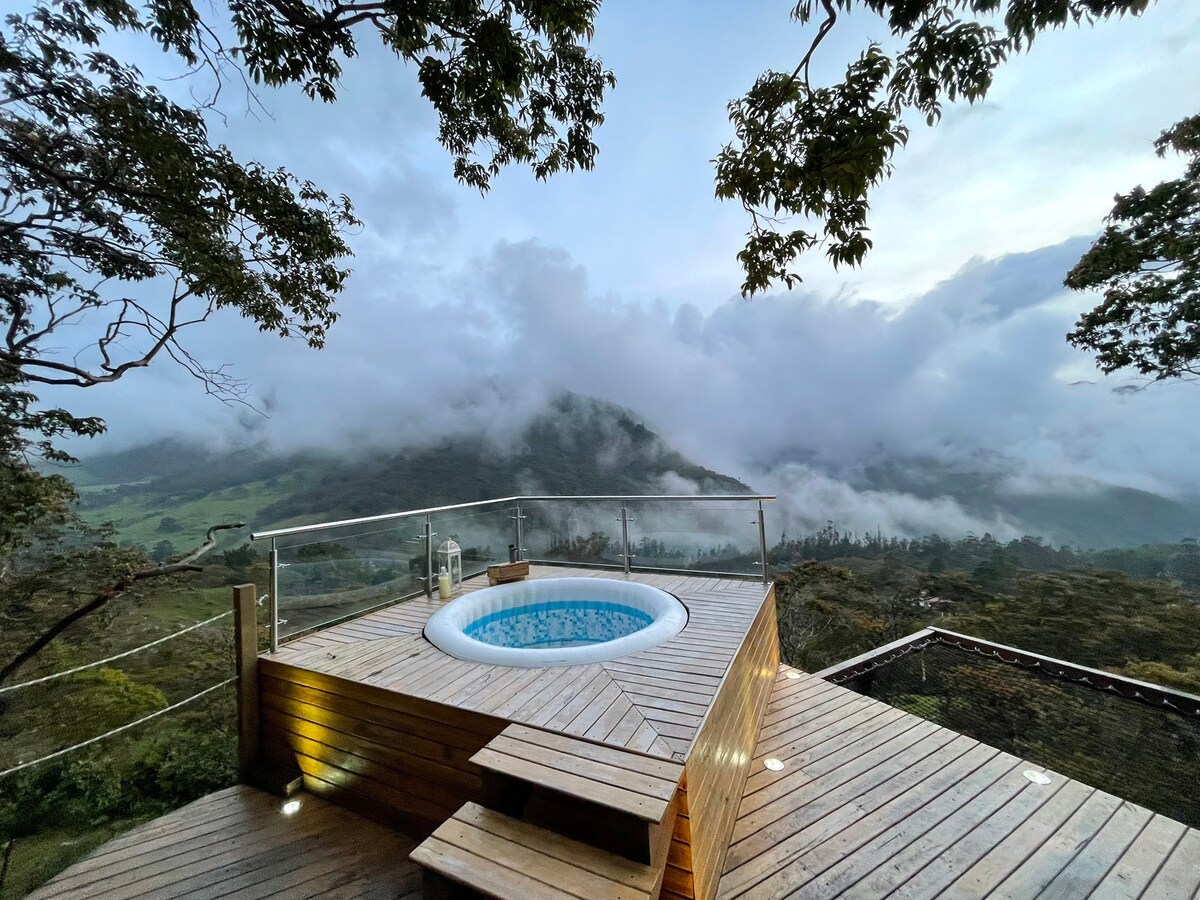 A wooden deck is featured, equipped with a circular jacuzzi that gently blends with the landscape. A lush view of rolling hills and layers of mist creates a serene atmosphere, while trees provide a sense of privacy around the deck.