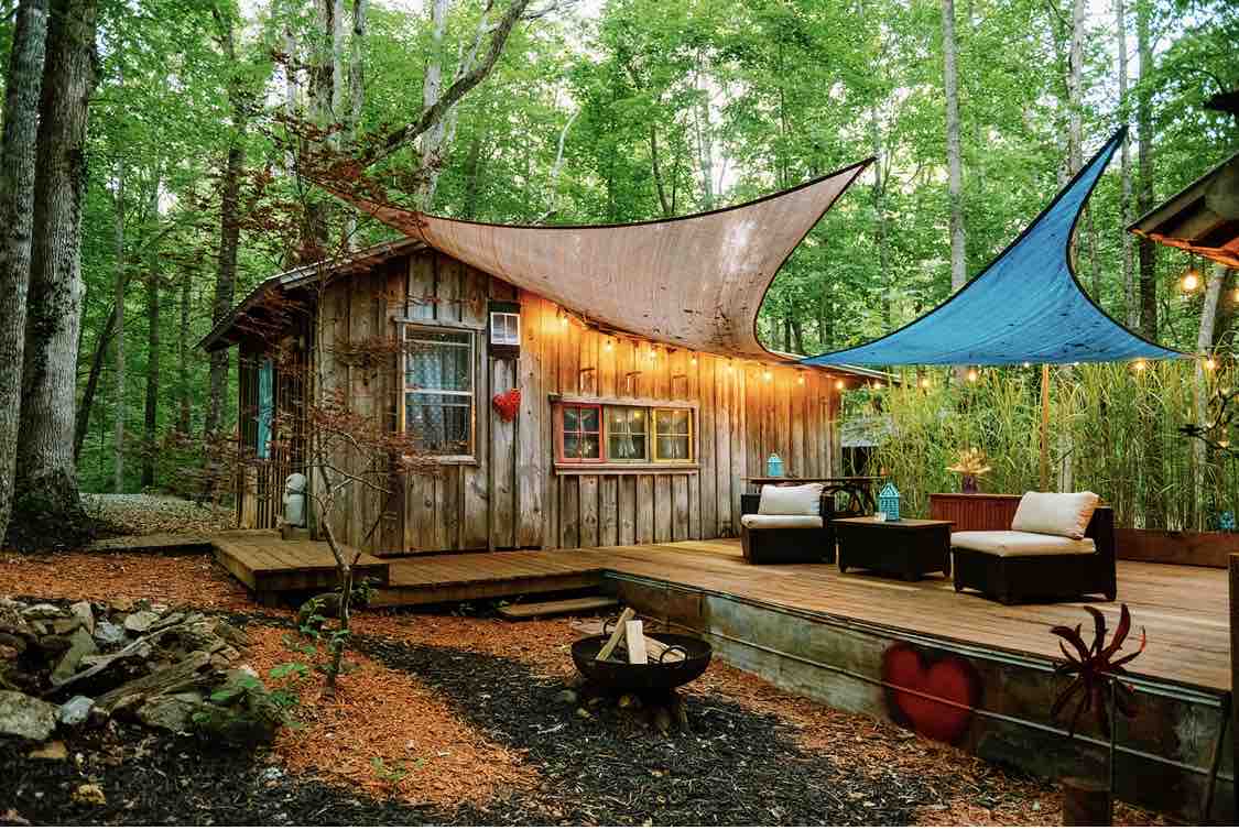 A rustic cabin nestled among lush greenery features a wooden deck complemented by stylish outdoor seating. Colorful fabric sails provide shade, while warm lights enhance the inviting atmosphere. A fire pit area surrounded by natural stone adds to the serene outdoor setting.