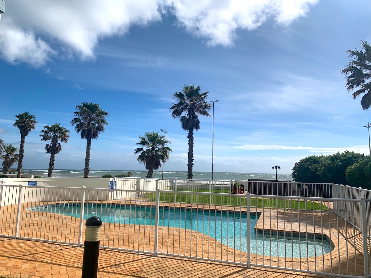 A fenced swimming pool is situated in the foreground, bordered by palm trees. The serene ocean can be seen in the background under a clear blue sky, with a hint of clouds adding depth to the scene.