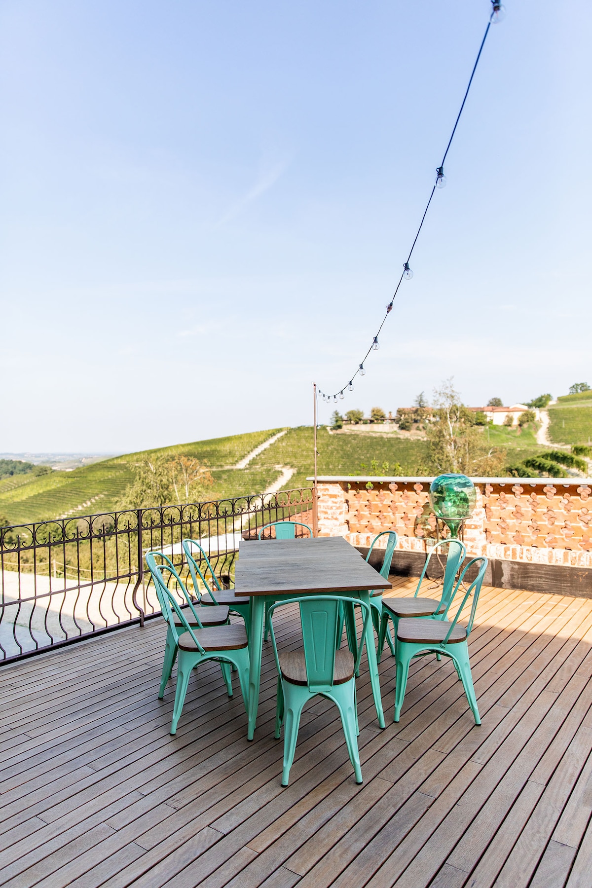 A terrace is shown, featuring a long dining table surrounded by six teal chairs. The area is enhanced by a subtle string of decorative lights overhead. A scenic view of rolling hills in the background adds to the tranquil setting.