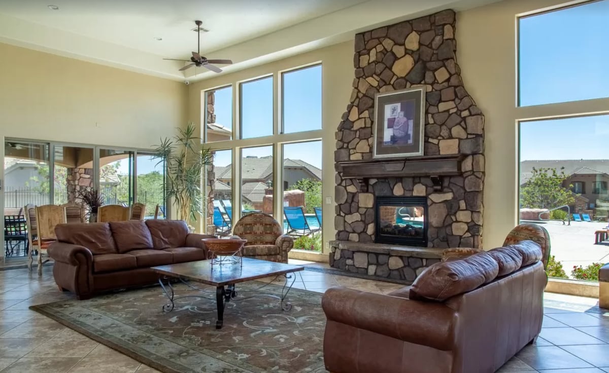 A spacious lounge area is highlighted by large windows allowing natural light to flood in. A stone fireplace serves as a focal point, surrounded by comfortable seating options including leather sofas and an elegant coffee table. Outdoor seating can be seen through the windows.