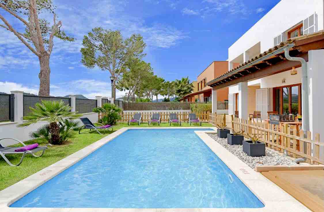 The outdoor area features a swimming pool surrounded by a spacious garden. Sun loungers are arranged on one side, while a wooden fence offers safety. The villa's exterior showcases large windows and a covered patio area designed for outdoor dining, framed by lush greenery.