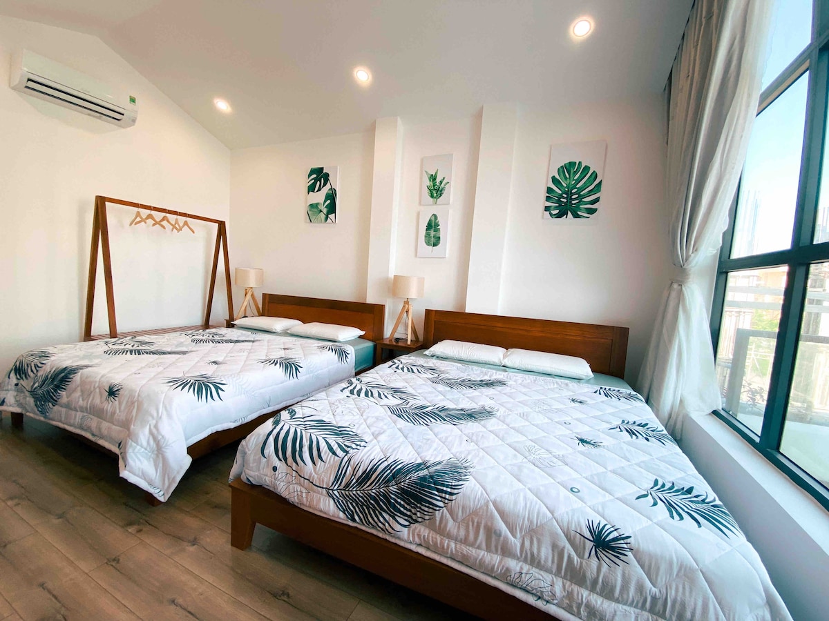 A bright and airy guest room features two spacious beds adorned with leaf-patterned bedding. Large windows provide natural light, highlighting the minimalist decor and botanical wall art. The room is equipped with air conditioning, ensuring a comfortable stay.