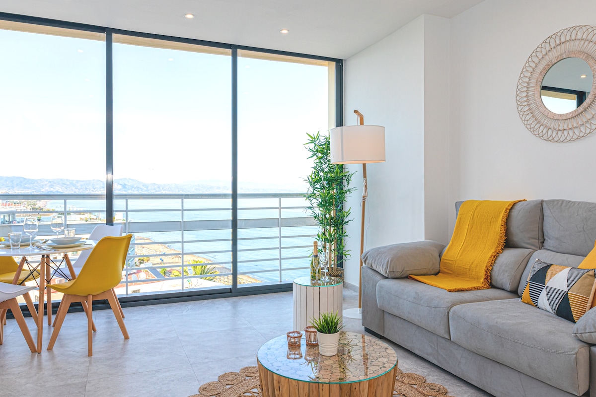 A light-filled living area features a spacious gray sofa adorned with a decorative throw. A circular coffee table sits in front, complemented by a small potted plant. Large glass windows provide panoramic sea views, while a dining table for four offers a scenic dining experience.