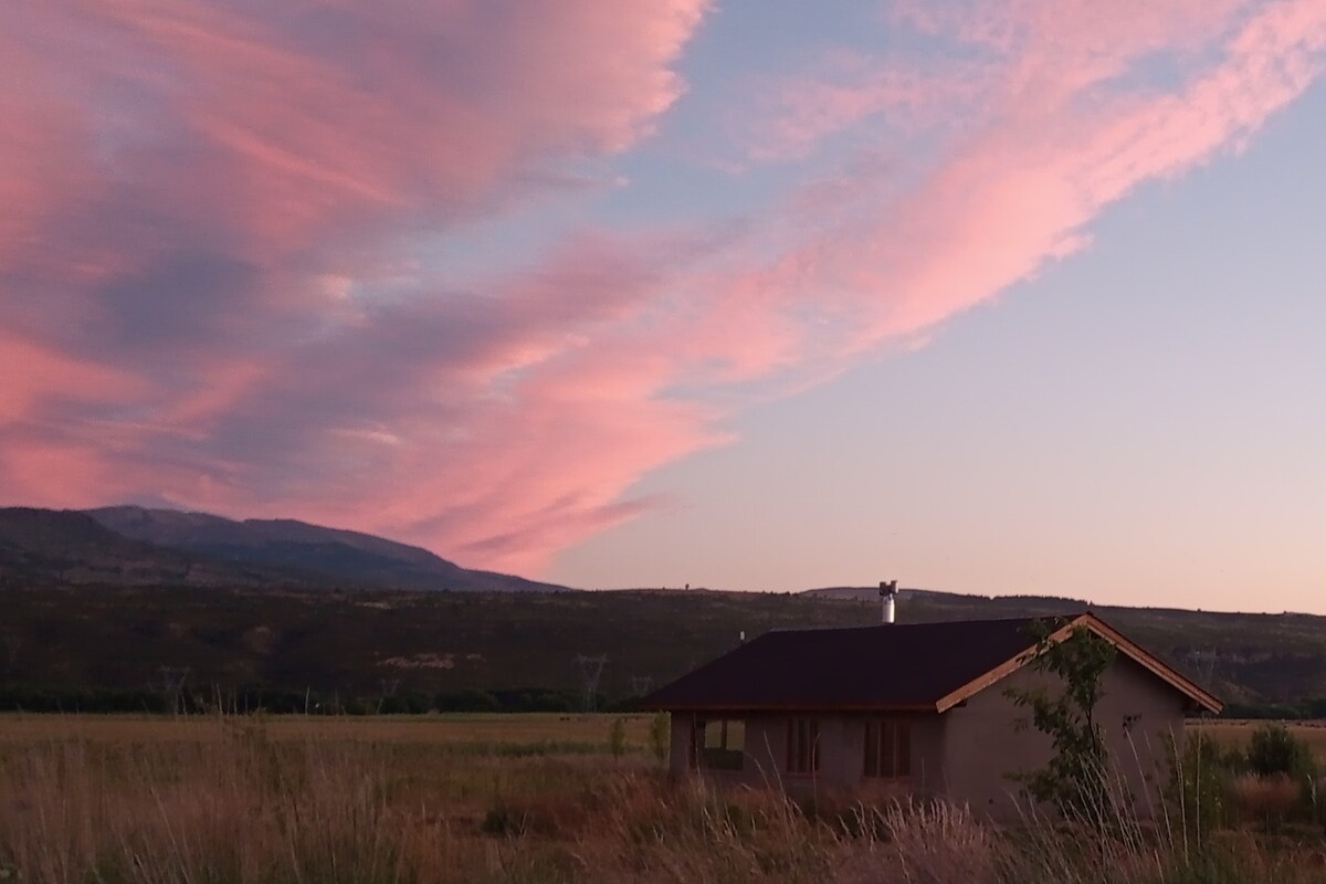 The image depicts a modest house constructed with natural materials, set against a backdrop of mountains and colorful clouds at sunset. Expansive fields surround the home, providing a serene rural environment. Soft grasses and plants frame the foreground, enhancing the peaceful landscape.