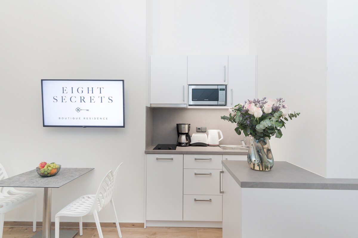A modern kitchen area is featured with sleek white cabinetry and a countertop. A Smart TV mounted on the wall displays the logo of 'EIGHT SECRETS – BOUTIQUE RESIDENCE'. A dining table with two white chairs is set, accompanied by a vase of fresh flowers and a bowl of colorful fruit.