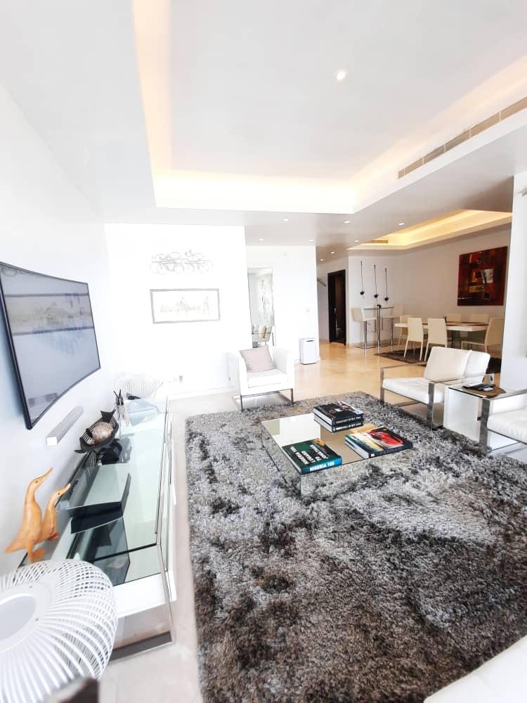 A spacious living area features modern furniture, including a sectional sofa and sleek coffee table. The soft, textured rug anchors the space, while a large flat-screen TV is mounted on the wall. Natural light enhances the inviting atmosphere, complemented by warm ceiling lighting.