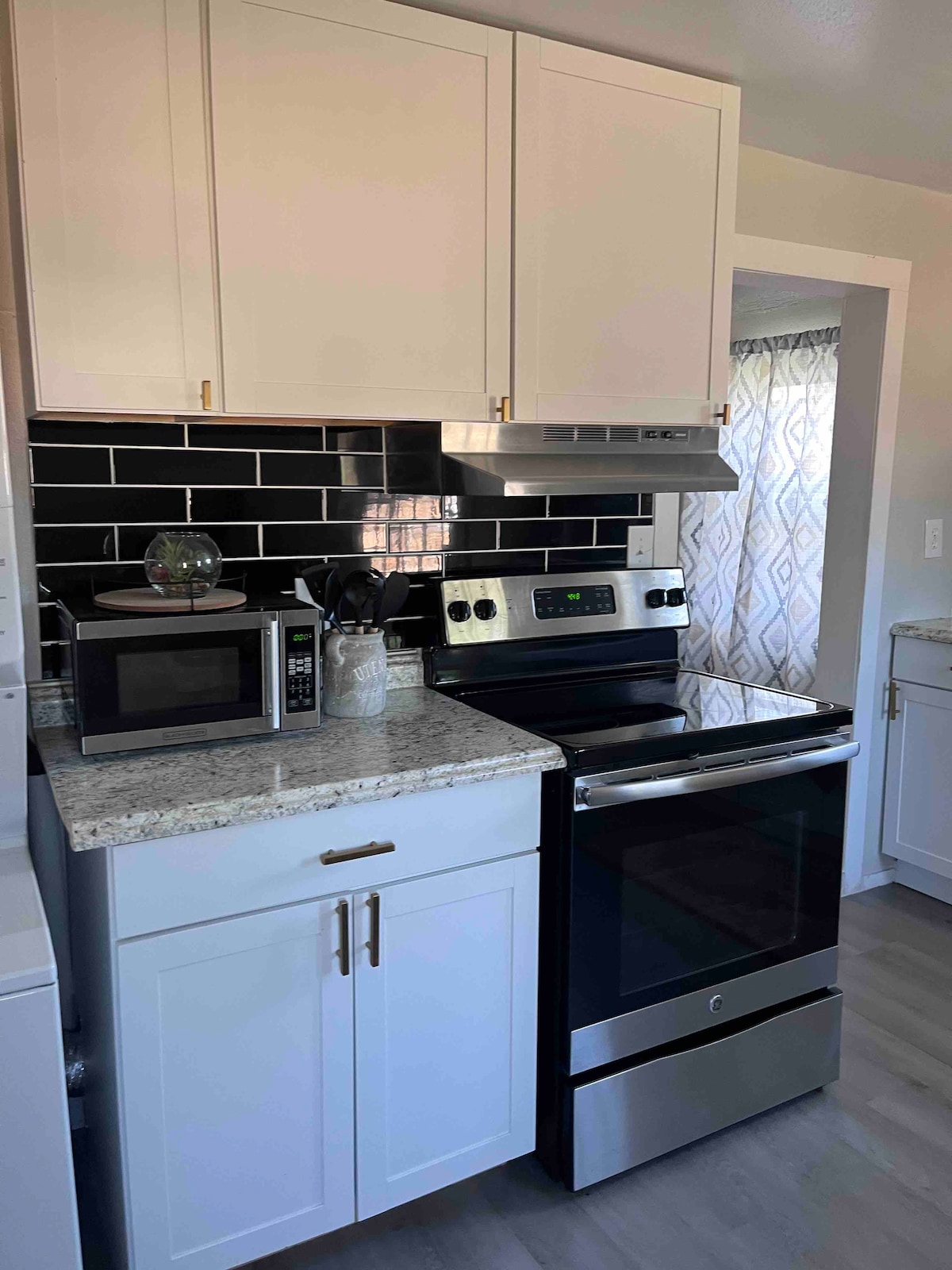 The kitchen features modern white cabinetry paired with a contrasting black tile backsplash. A stainless steel range and a microwave are present, with ample counter space visible, including a granite surface. Natural light enters through a window dressed with patterned fabric.