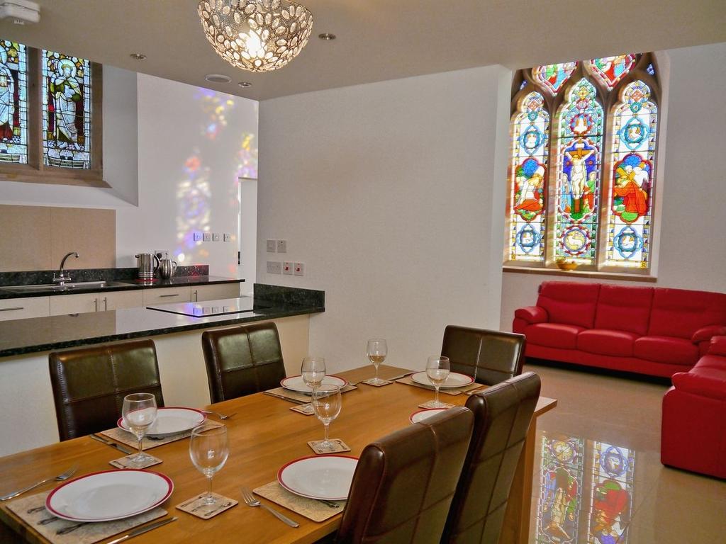 A spacious dining area is highlighted by a wooden table set for six, accompanied by brown leather chairs. The contemporary kitchen is visible in the background, while vibrant stained glass windows illuminate the room with colorful reflections. A red sofa adds a pop of colour to the space.