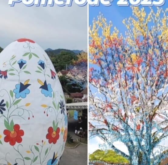 A large, decorative egg features colorful floral patterns, situated in an outdoor setting. Next to it, a vibrant tree is adorned with multicolored lights, creating a striking visual contrast against the natural backdrop.