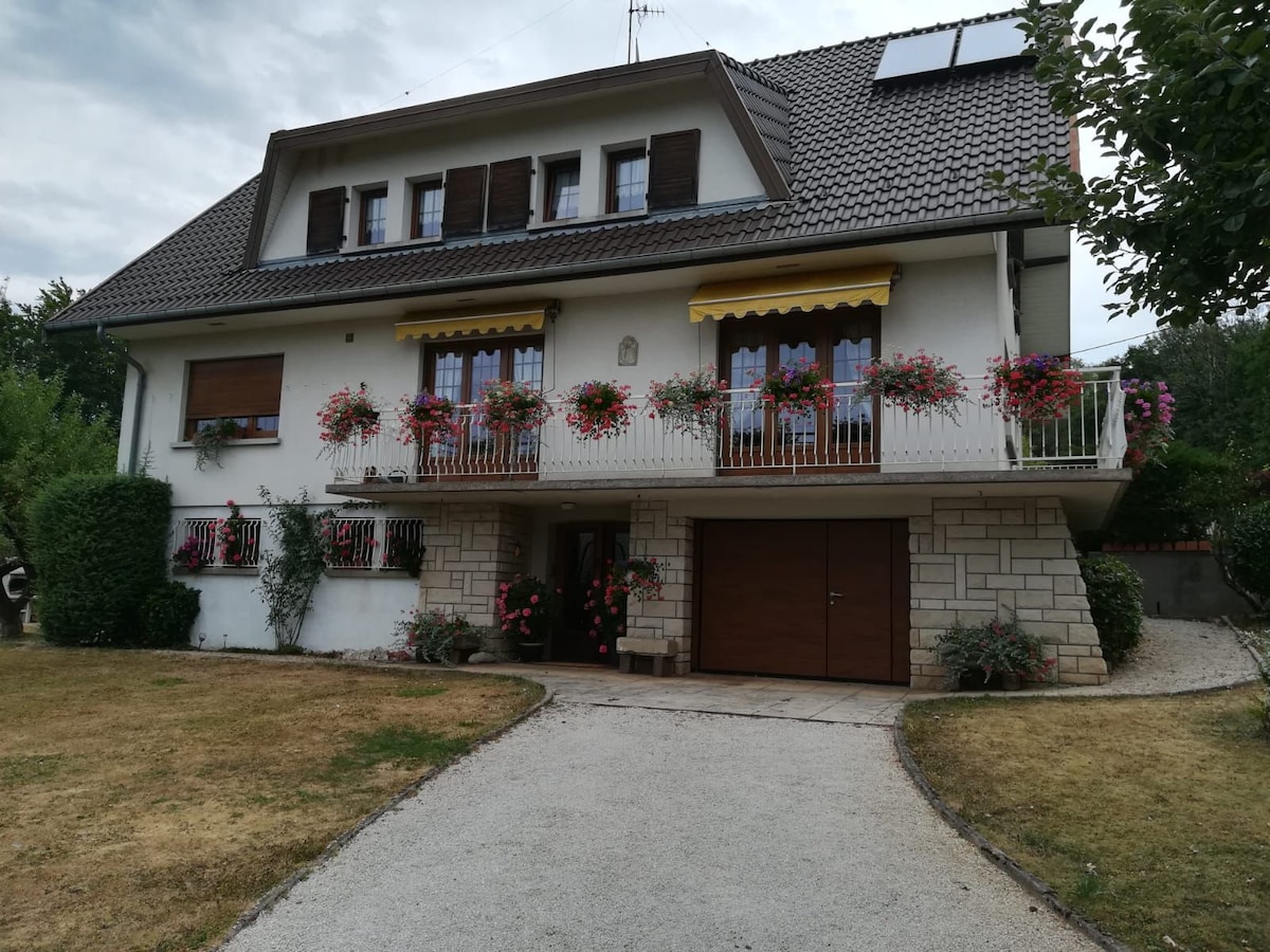 The two-story house is framed by a well-maintained garden, featuring a gravel driveway leading to a garage. Flower boxes hang from the balconies, adding vibrant color to the white facade. Overhanging awnings provide shaded areas on the upper deck.