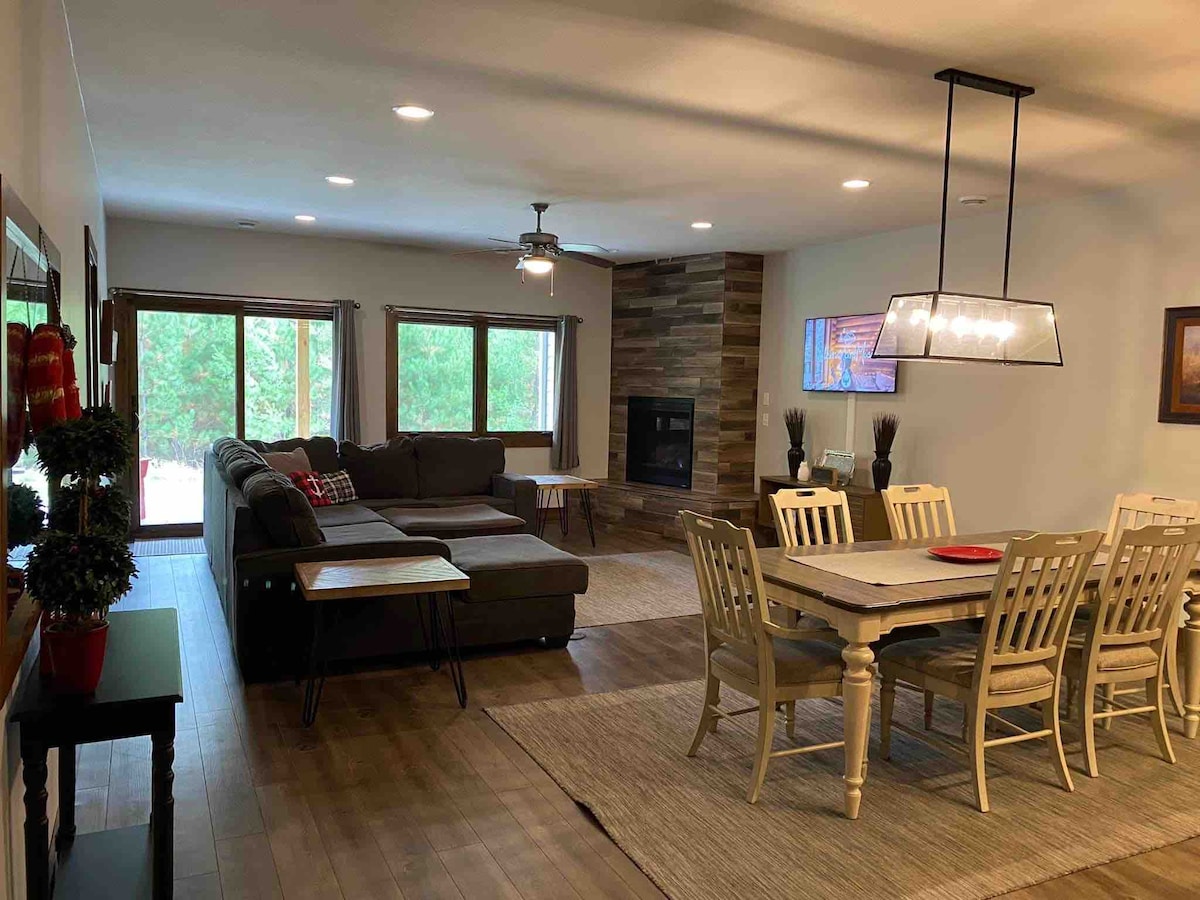 A spacious living area features a comfortable sectional sofa arranged around a coffee table. A dining table with six chairs is positioned nearby. Natural light floods the space through large windows, highlighting the stone accent wall and a flat-screen television mounted above a modern fireplace.