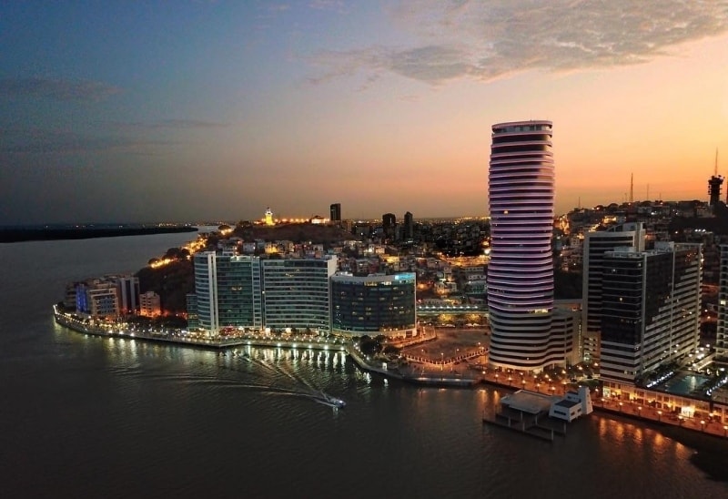 The cityscape is captured at dusk, featuring modern high-rise buildings along the waterfront. Soft lights illuminate the structures as the sun sets, reflecting warm hues on the calm water. A hint of distant hills and a clear sky complete the serene evening view.