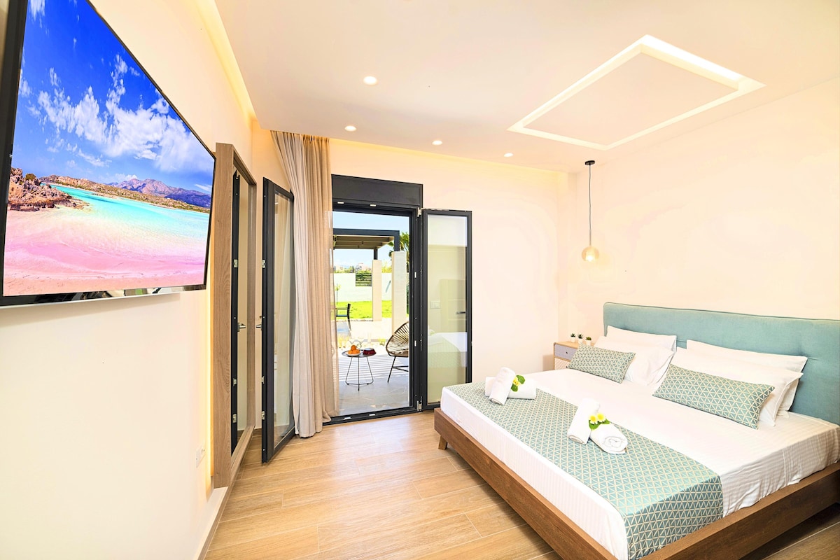 A bright bedroom features a spacious bed with light bedding and decorative pillows. A wall-mounted TV is positioned opposite the bed. Large glass doors open to a private terrace, allowing natural light and offering views of the outdoor area.