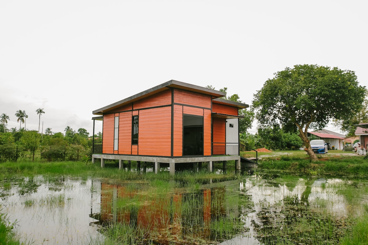 A striking cottage is situated on stilts above a reflective body of water, surrounded by lush greenery. Its vibrant orange exterior contrasts with the natural landscape, while palm trees and a nearby vehicle are visible in the background.