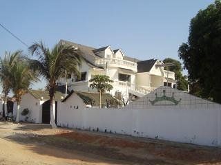 The exterior of a multi-level building is shown, featuring a white facade complemented by a spacious balcony on the second floor. Palm trees are visible nearby, providing greenery to the scene. The property is enclosed by a white wall with an entrance gate.