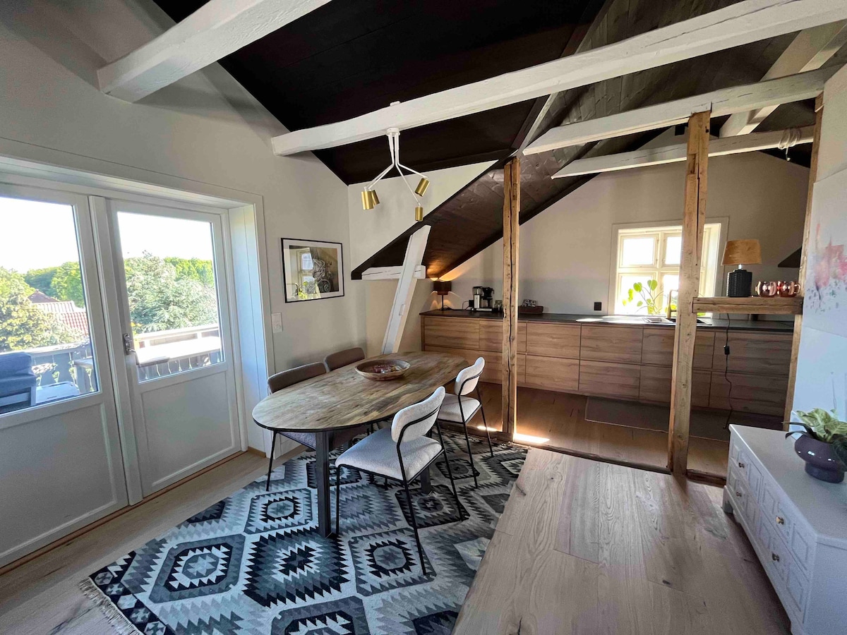 A dining area features a round wooden table surrounded by five chairs, set against a patterned rug. Natural light enters through large windows, illuminating the space, while exposed beams and wooden accents add a rustic touch to the modern kitchen area in the background.