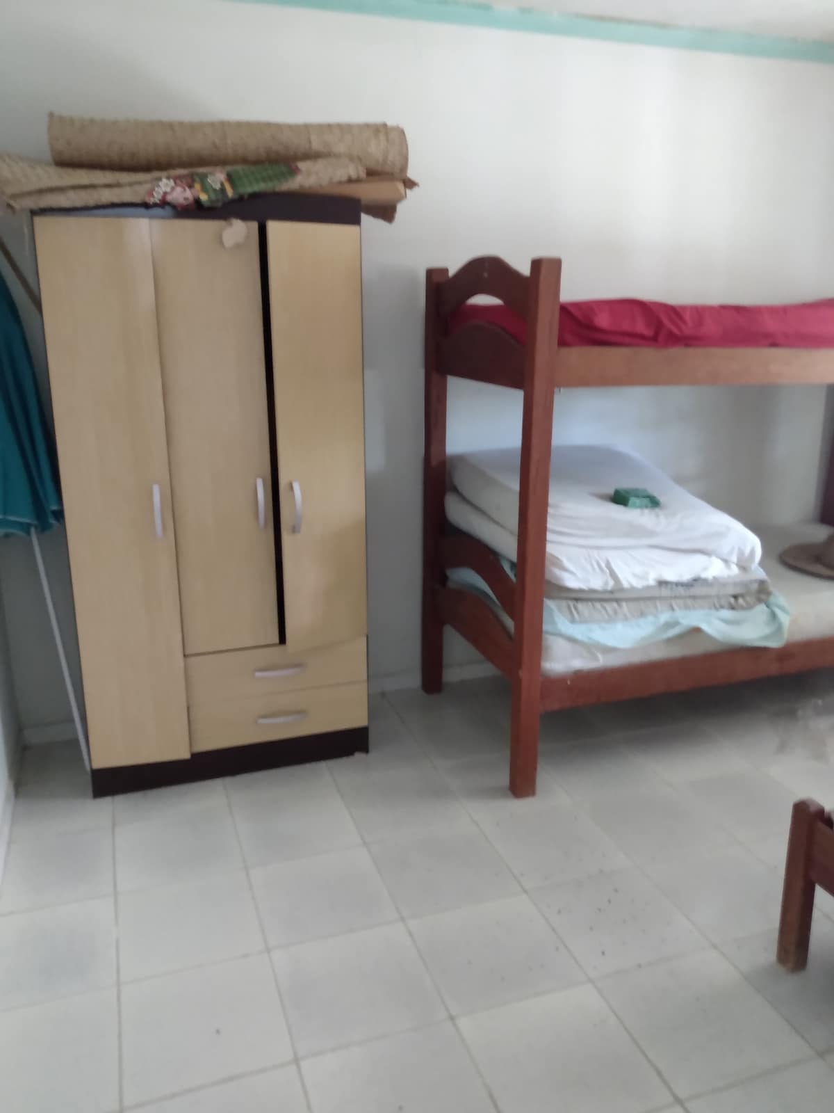 The image captures a simple room featuring wooden bunk beds with red bedding. A spacious cabinet stands against the wall, and a few mattresses are stacked nearby. The floor is composed of neutral tiles, contributing to an open and functional layout.