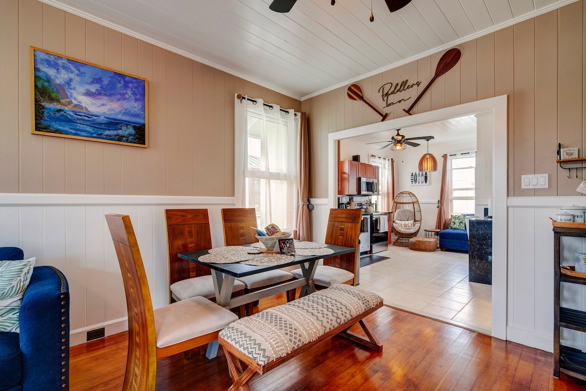 A dining area features a wooden table accompanied by several chairs, with a cozy bench positioned nearby. Natural light filters through large windows, and a painting of the ocean adorns the wall. A view of the open kitchen is visible in the background.