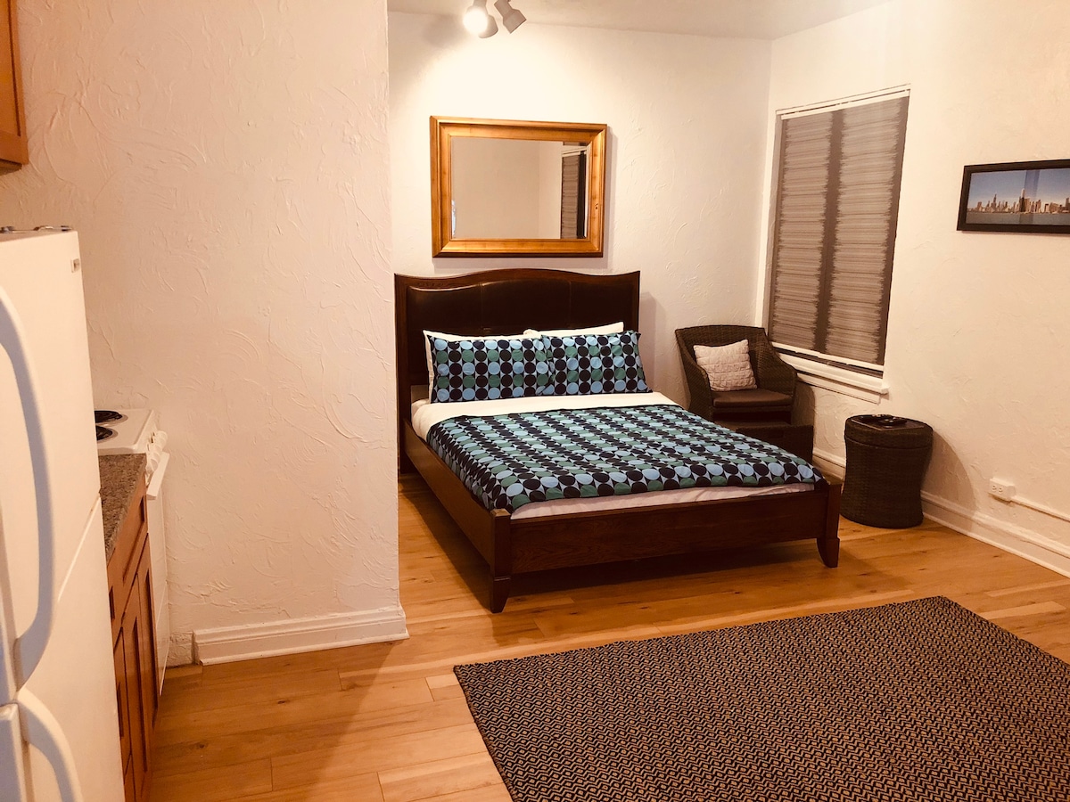 A studio space features a spacious bed with a patterned blanket, complemented by a decorative mirror above the headboard. A small seating area with a chair and woven basket is situated beside a window adorned with privacy blinds. The floor is covered with a textured area rug.