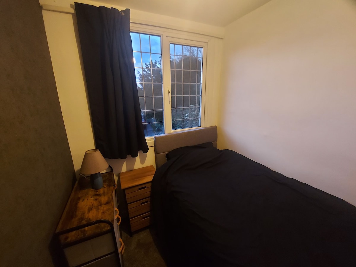 A cozy bedroom is presented, featuring a single bed with dark bedding. A wooden nightstand is positioned beside the bed, accompanied by a small lamp. A large window with dark curtains allows natural light to enter, while a soft carpet complements the room's decor.