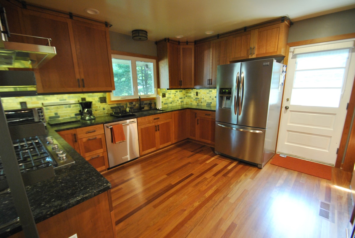 The newly remodeled kitchen features warm wooden cabinets and sleek granite countertops. A stainless steel refrigerator and modern appliances are visible, with green and yellow mosaic tiles adding a splash of color. Natural light enters through the window above the sink, enhancing the clean and functional layout.