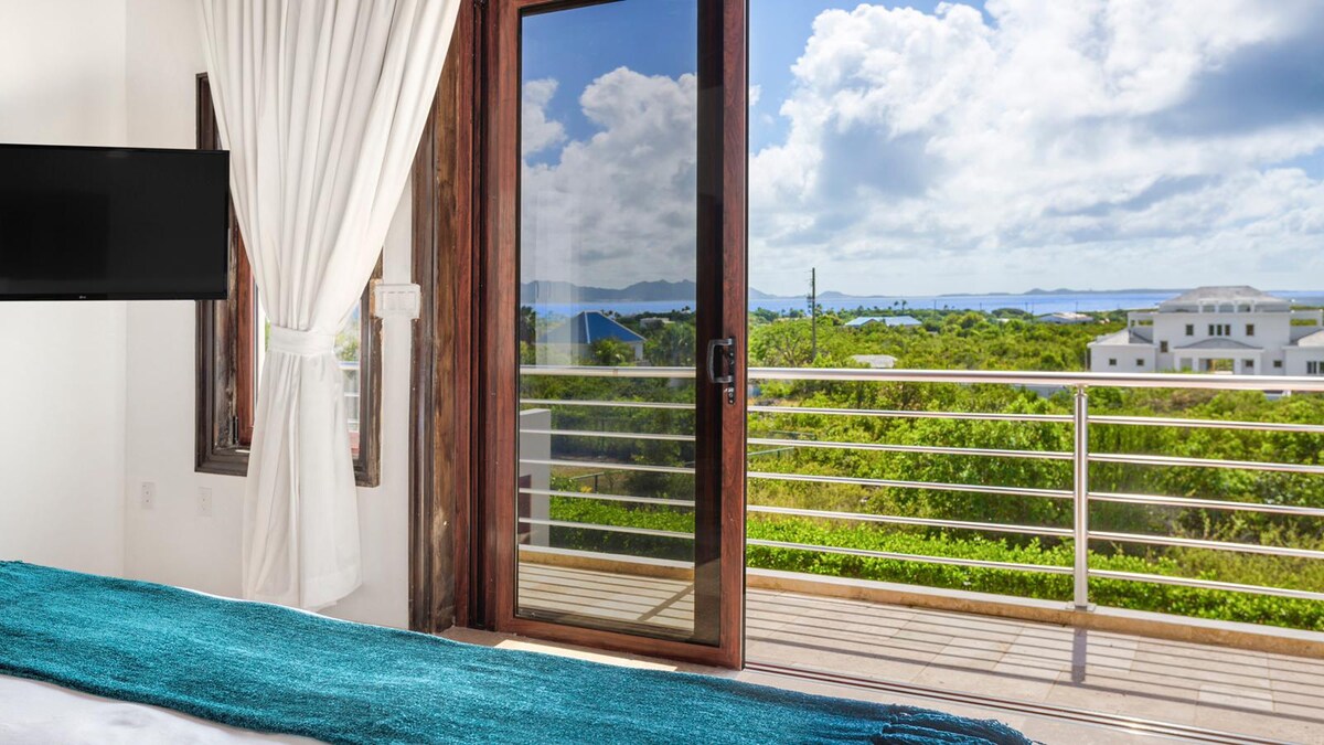 A view from the master suite showcases expansive glass doors that open to a private balcony. Lush green landscapes and distant blue waters of St. Maarten are visible under a partly cloudy sky. The room features light curtains and a turquoise accent draped over the bed.