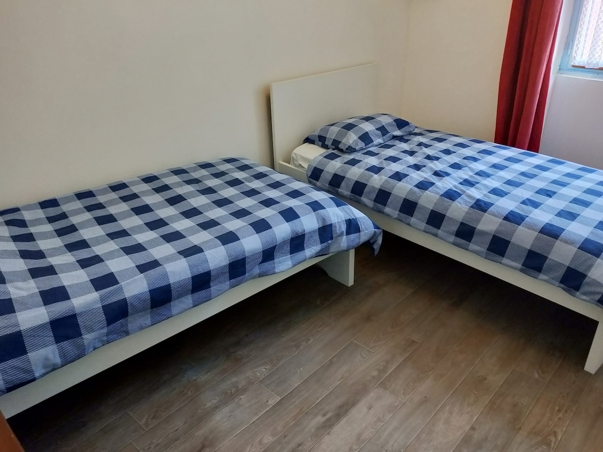The room features two twin beds positioned side by side, each covered with blue and white checkered bedding. Light-colored walls and wood flooring create a neutral backdrop. A window with red curtains offers natural light, enhancing the room's brightness.
