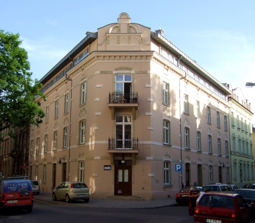 A historic, elegantly restored building from 1905 is depicted with a light-colored facade and intricate architectural details. Balconies are visible on the upper floors, and trees are seen nearby, contributing to the pleasant urban environment.
