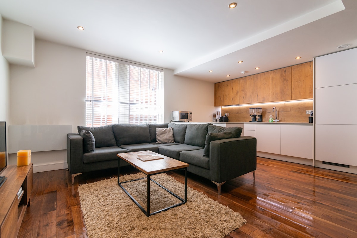 A modern living area is presented with a dark sectional sofa and a coffee table atop a light area rug. Natural light enters through a large window, illuminating the adjoining kitchen with wooden cabinetry and stainless-steel appliances. The open layout creates a welcoming space for relaxation.