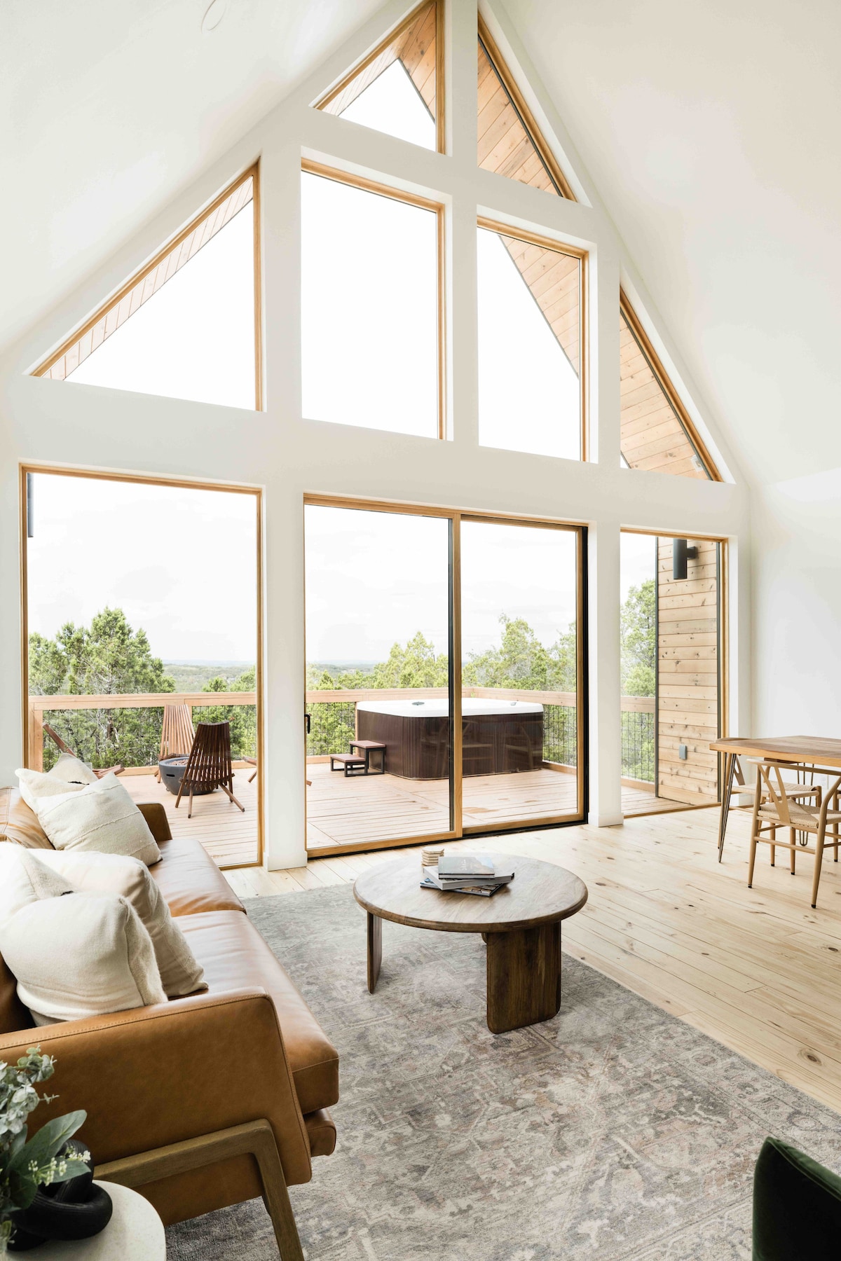 A spacious living area features a leather sofa, round table, and large glass windows that offer views of the surrounding landscape. Natural light floods the space, highlighting a modern, minimalist design with wood accents. A door leads to an outdoor deck with a hot tub.