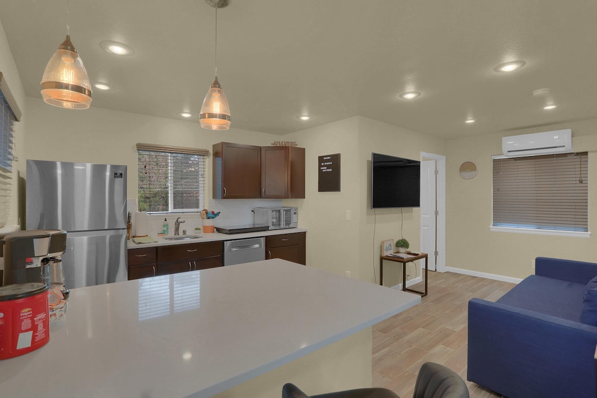 The kitchen features modern appliances, including a refrigerator and coffee maker. A spacious island with a light quartz countertop serves as a dining area. Illuminated pendant lights hang overhead, and a comfortable seating area is visible across from the kitchen.