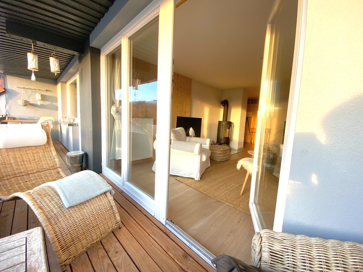 A comfortable living space is visible through large glass doors, allowing natural light to fill the area. A cozy seating arrangement with a neutral color palette is located within, complemented by a light-colored rug and a wood stove. Outdoor seating is present on the balcony.