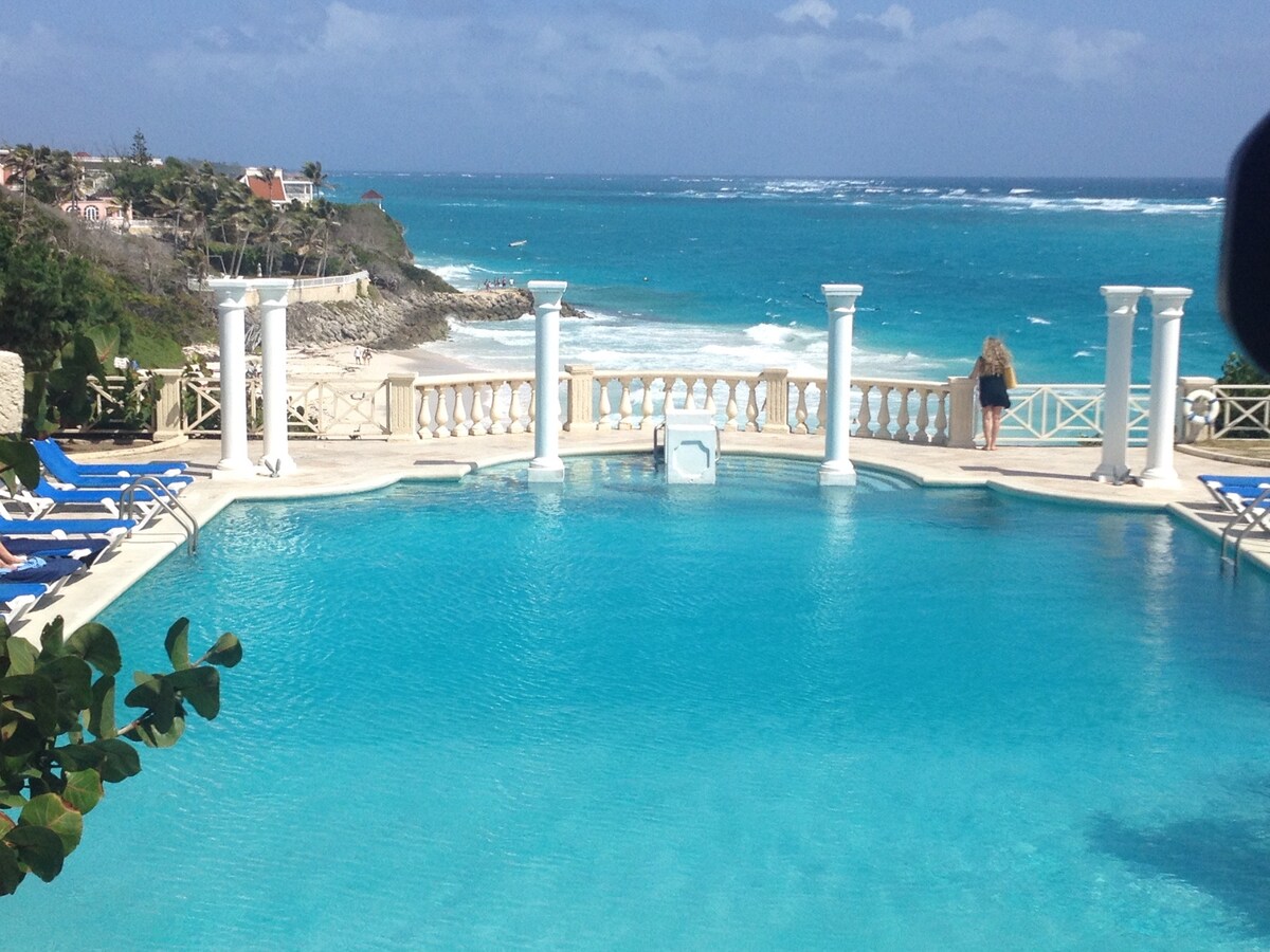 A large outdoor pool is framed by elegant white columns, leading to a breathtaking view of the Atlantic Ocean. Sun loungers are positioned around the pool, offering a relaxing space to enjoy the scenery. The sky displays a mix of clouds and blue, reflecting the inviting water.