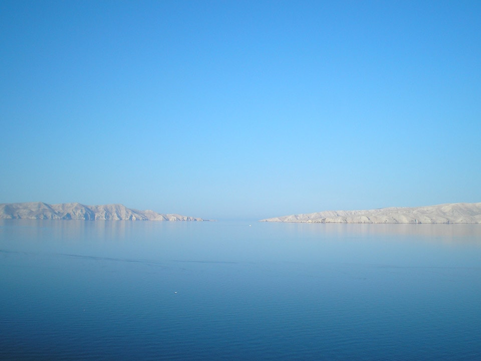 A panoramic view of calm waters reflecting a clear blue sky, framed by distant mountains that gently slope toward the sea. The tranquility of the scene suggests an inviting atmosphere, ideal for relaxation and enjoyment of nature.