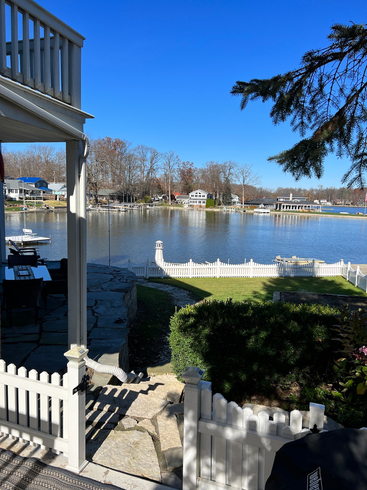 A view of the calm lake is framed by a white picket fence, with grassy areas leading down to the water. The scene includes nearby homes and boats, complemented by clear blue skies and the natural greenery surrounding the area.