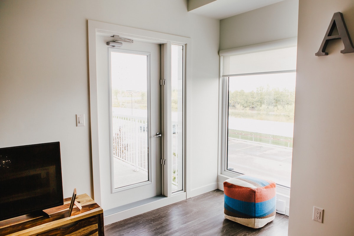 A corner of a modern living space is shown, featuring a large window with a view and a door leading to a balcony. A colorful pouf is positioned on the floor, and a television is placed on a wooden console nearby.