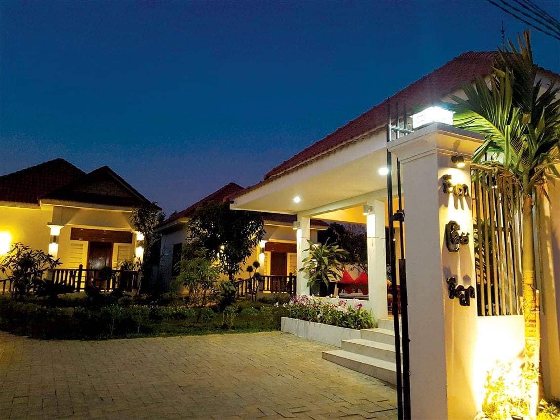 The villa exterior is warmly lit, showcasing a modern design and well-maintained landscaping. The entrance features a stone pathway leading to the main doors, surrounded by lush greenery and colorful flower beds, creating a welcoming ambiance during the evening.