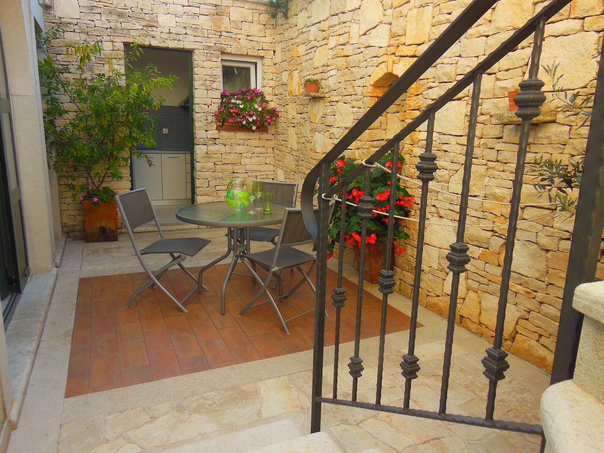 A charming courtyard features a circular glass table surrounded by four metal chairs. Lush greenery and colorful flowers are situated against a rustic stone wall, creating a serene outdoor space. Natural light softly illuminates the area, emphasizing the tranquil setting.