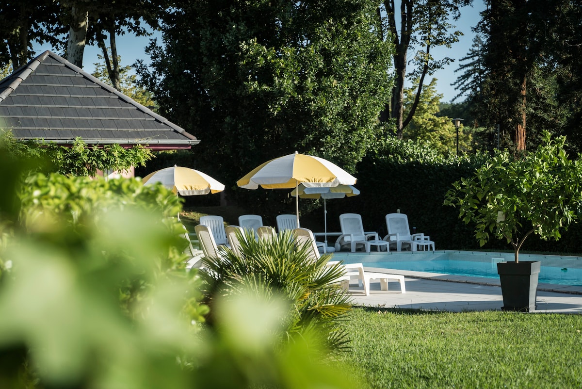 A heated pool area is surrounded by lush green shrubs and trees. White lounge chairs are positioned under yellow and white umbrellas, offering shaded seating by the water. The outdoor space is complemented by manicured grass and a nearby decorative plant.