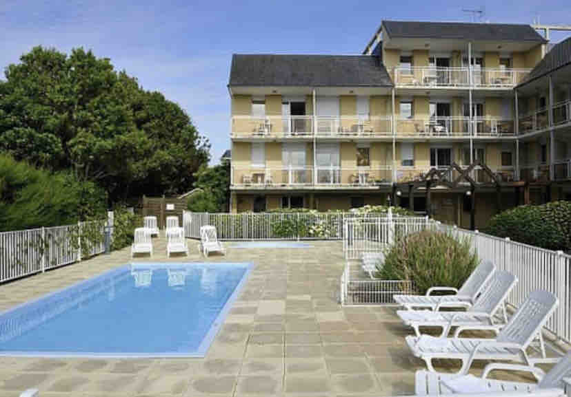 An outdoor area features a swimming pool surrounded by tiled flooring and a fence. Lounge chairs are arranged nearby, with greenery visible in the background. The multi-story building has balconies overlooking the pool area, creating a peaceful setting for relaxation.