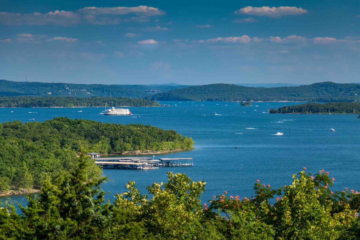 A panoramic view of a tranquil lake is captured, featuring numerous boats gently navigating the water. Lush green trees frame the scene, while distant hills rise against a clear blue sky with soft, scattered clouds.