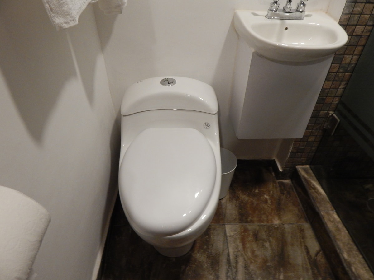 A clean and modern bathroom setting is displayed, featuring a white toilet and a compact sink mounted on the wall. The floor is covered in tiles, and a small trash can is positioned beside the toilet.