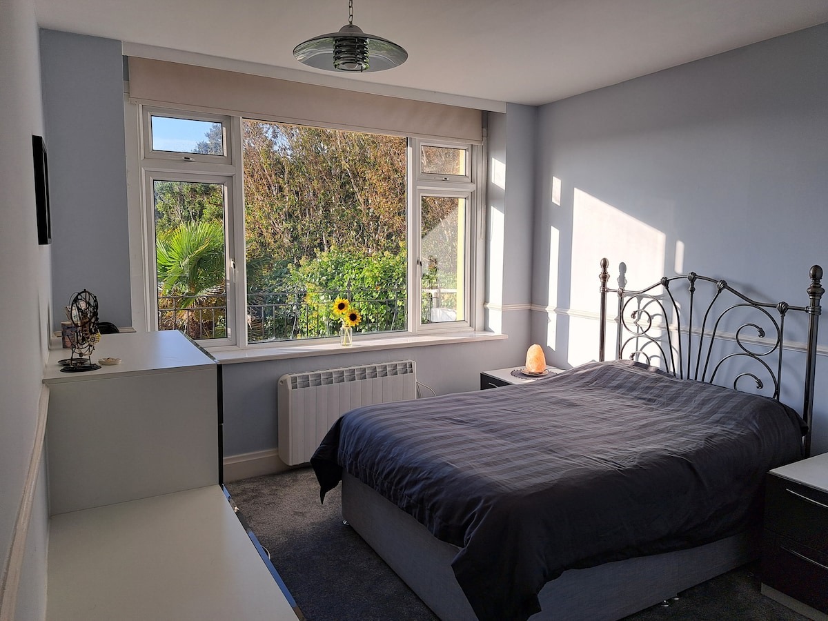 An ensuite bedroom features a spacious bed with a dark comforter, set against a light grey wall. A large window allows natural light to fill the room, showcasing garden views with palm trees. A bedside table and a low dresser provide functional touches.