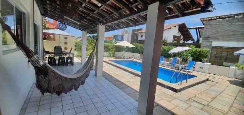 A covered patio area features a hammock and a dining set for six. The private pool is equipped with ladders and surrounded by lounge chairs and umbrellas, providing a relaxing outdoor space in a well-maintained garden.