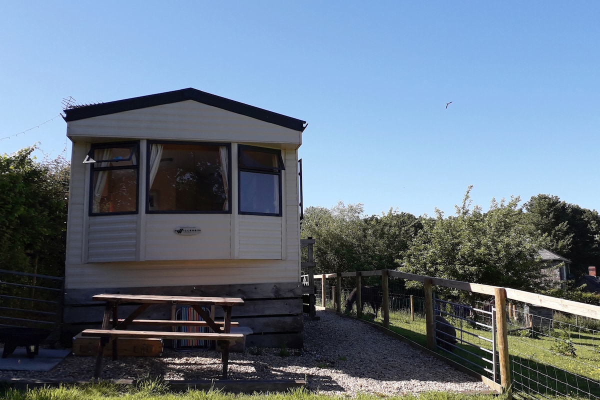 The holiday caravan is positioned on a grassy area, featuring large windows that allow for natural light. A picnic table is set outside, complemented by a wooden fence, with lush greenery visible in the background under a clear blue sky.