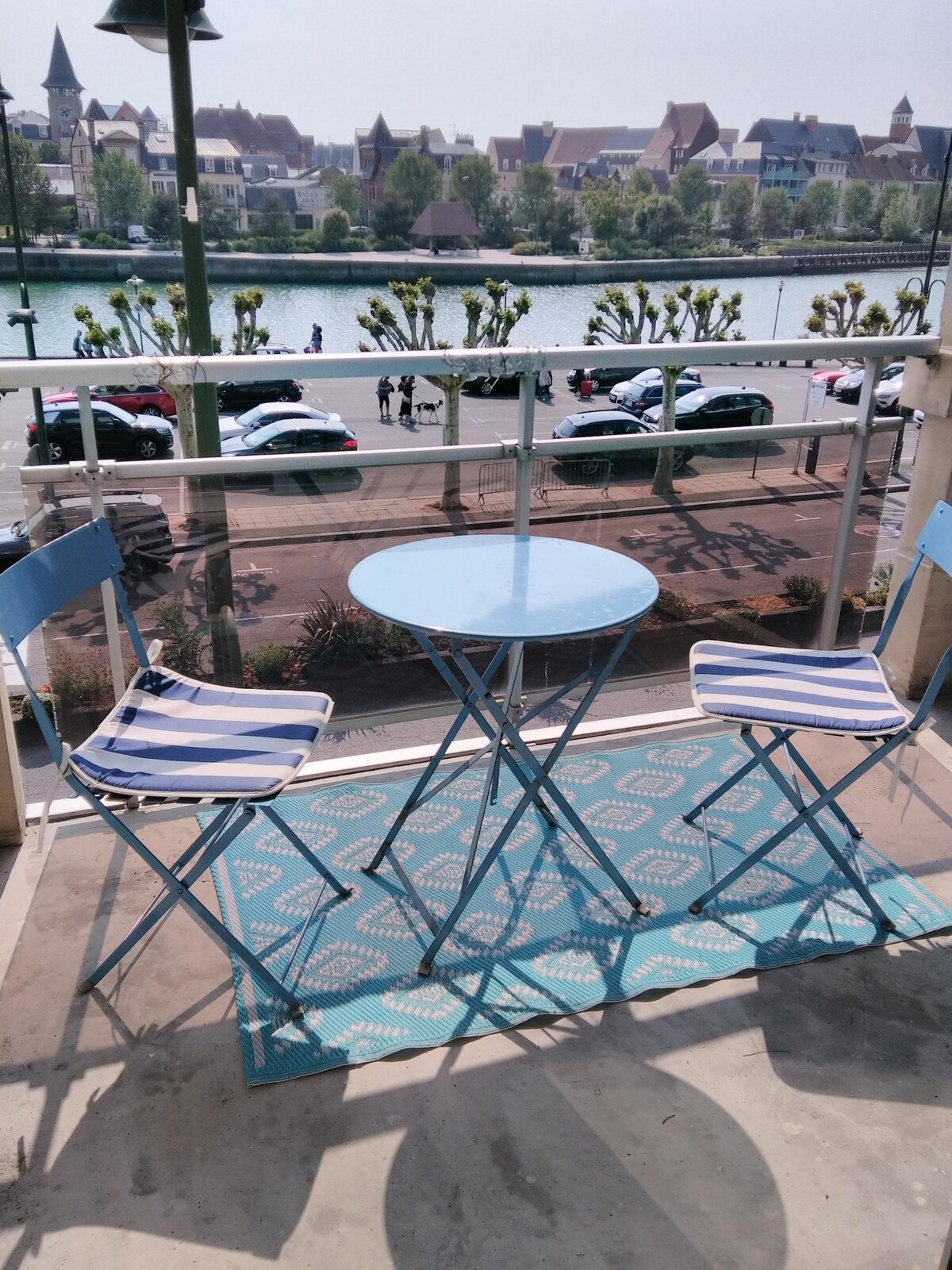 A small balcony area is featured with a round blue table and two striped folding chairs, set on a patterned rug. A scenic view of the port and surrounding architecture is visible in the background, enhancing the space's appeal for outdoor relaxation.