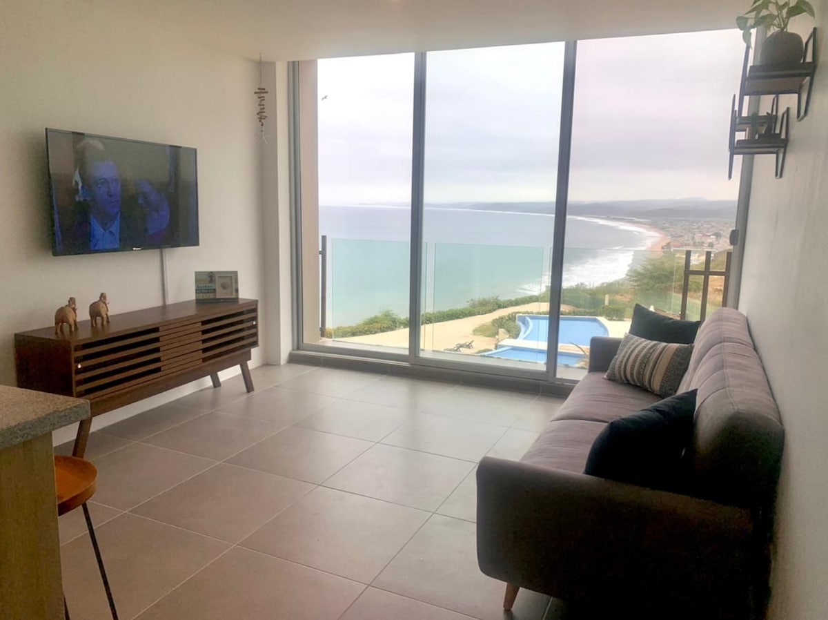 A modern living area is shown, featuring a large window that offers expansive views of the coastline. A gray sofa with accent pillows is positioned in front of a low wooden cabinet with decorative elements, and a flat-screen television is mounted on the wall.